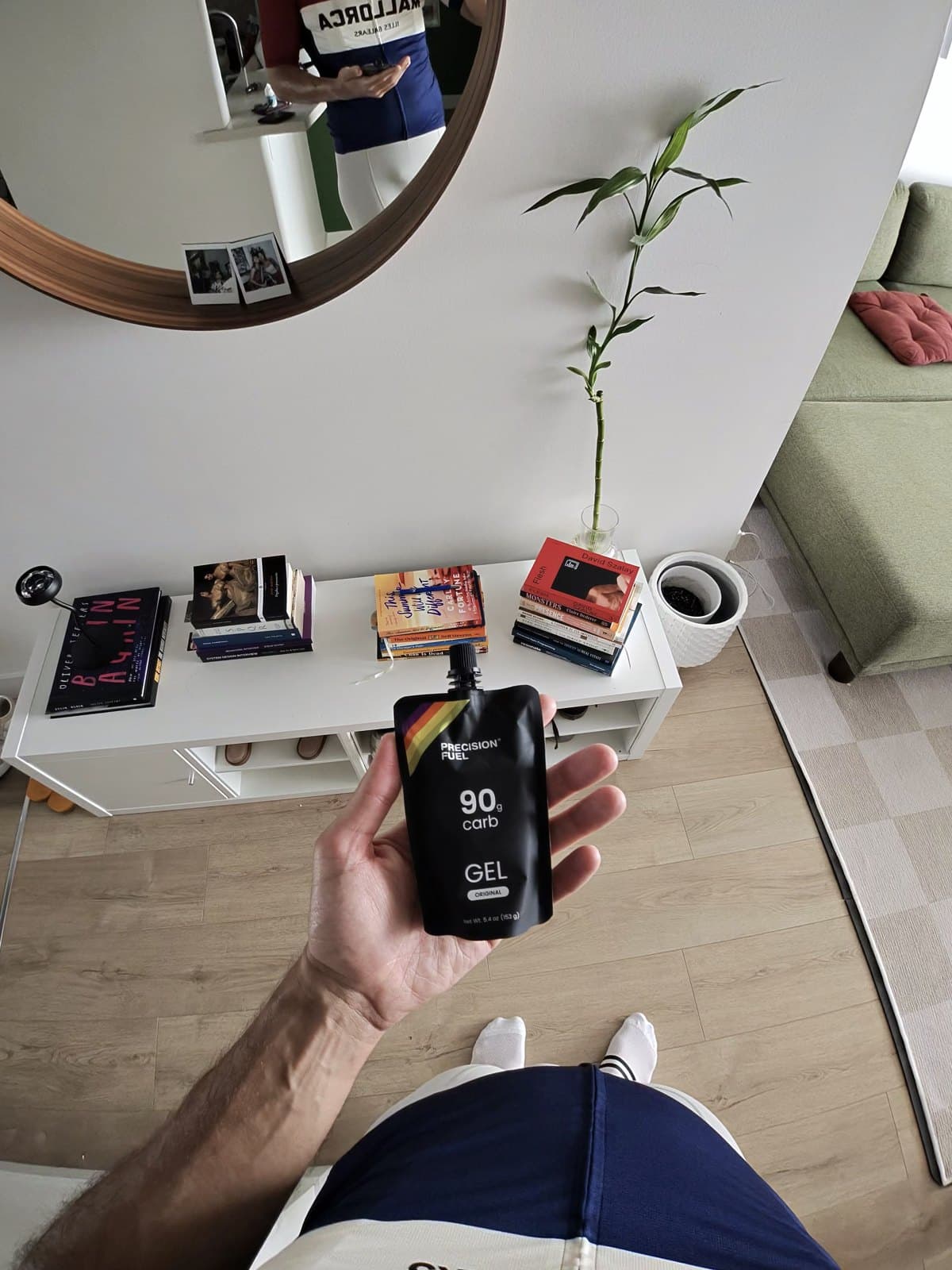 Lucas Calegari holding a Precision Fuel 90g carb gel flask before a long run, at home with a bookshelf and plant in the background