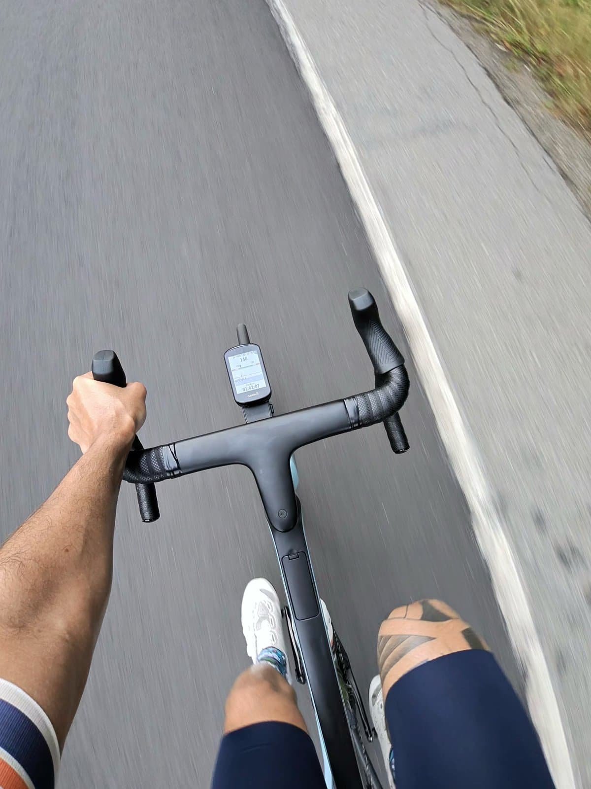 POV over the drop handlebars of the road bike on Sunday, one hand on the hood, bike computer mounted on the stem, road surface blurred with speed alongside the white shoulder line