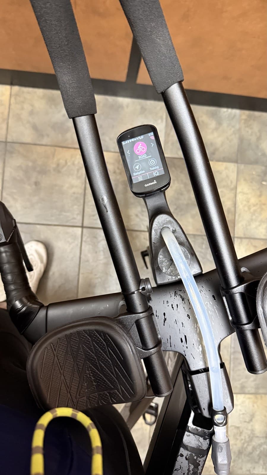 Top-down view of the tri bike cockpit — foam-padded aerobar extensions, a Garmin Edge bike computer showing the Road activity profile, and a hydration tube running down the centre, bike resting on a tiled floor