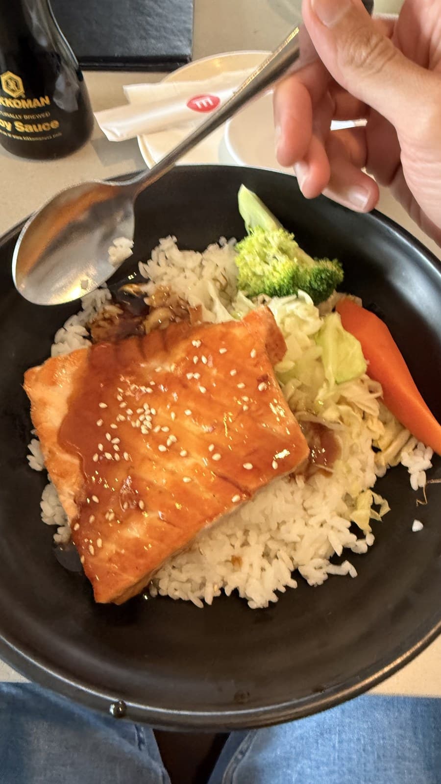 A bowl of teriyaki salmon over white rice with steamed broccoli, carrot and cabbage, a spoon and a bottle of Kikkoman soy sauce on the table