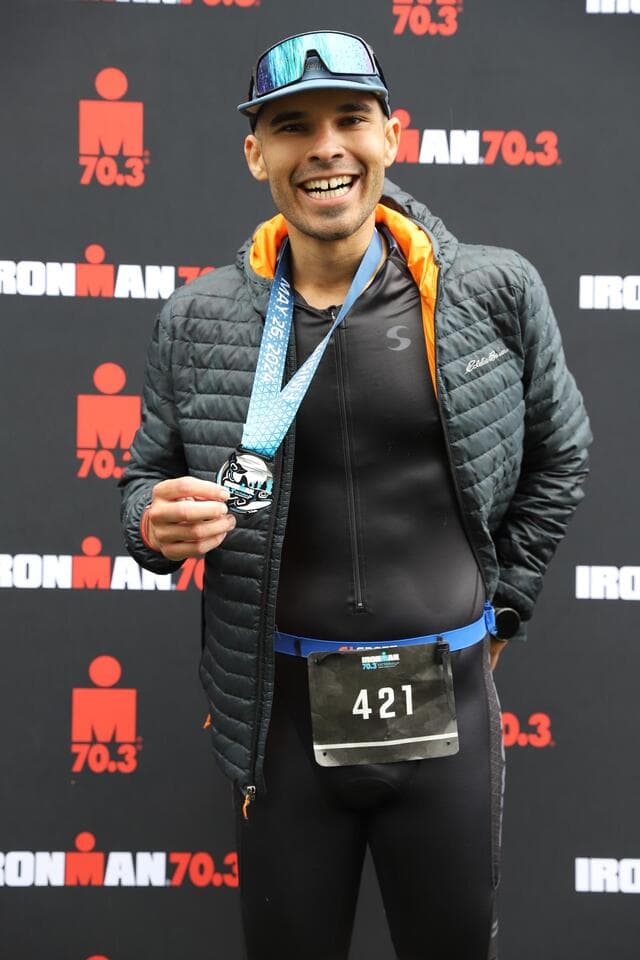 Lucas Calegari holding his Ironman 70.3 Victoria 2024 finisher medal in front of the IRONMAN 70.3 backdrop, race number 421
